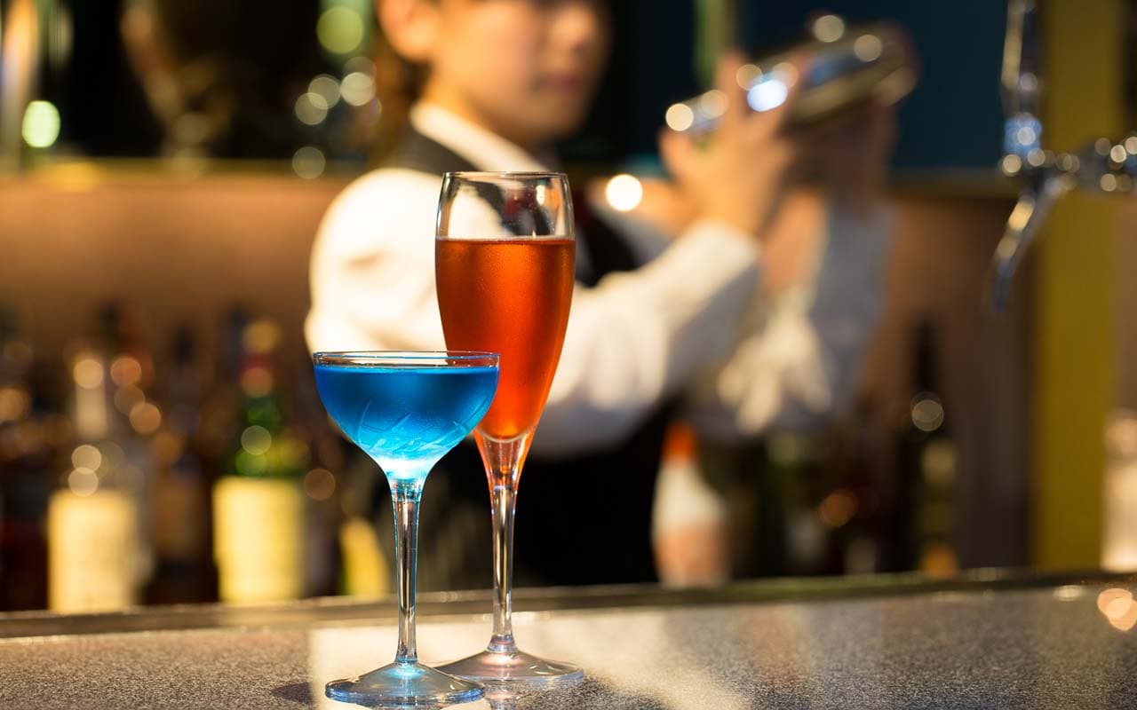 A bartender at Hotel Nikko Osaka preparing cocktails, with a focus on two vibrant drinks, one blue and one amber, displayed prominently on the bar counter, capturing the lively ambiance of the bar.