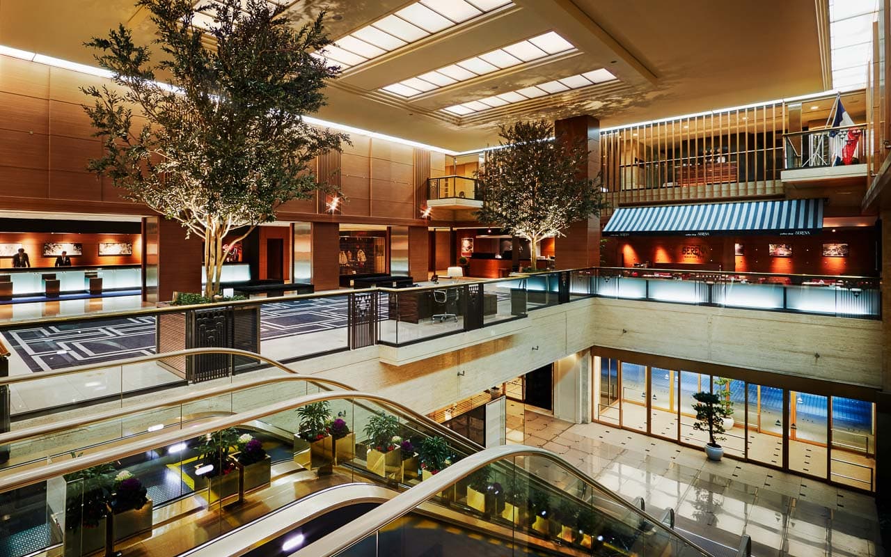 Opulent multi-level lobby of Hotel Nikko Osaka, featuring spacious interiors with elegant design elements, a large tree centrally positioned under natural light from the ceiling.