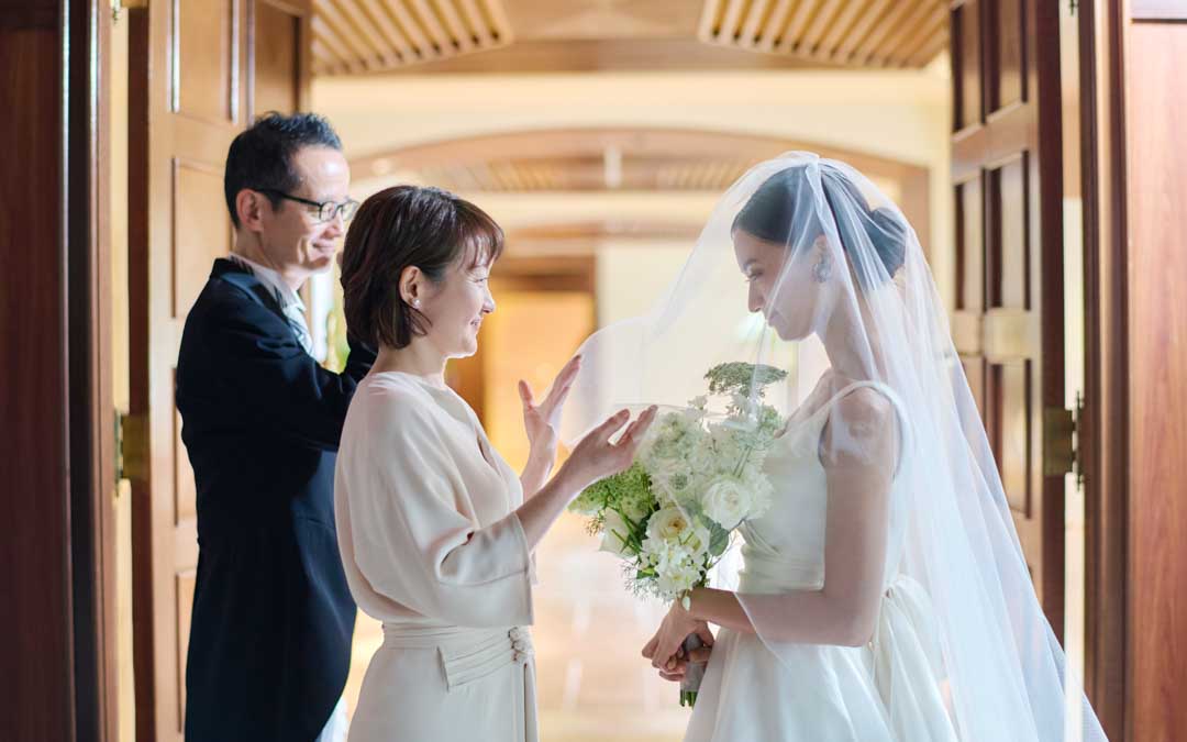 大きな扉の前に立ち、お互いに微笑み合う新婦と両親の写真。新婦はウエディングドレスとベールを身にまとい、両手に白いブーケを持っている。母親は軽く涙ぐみながら新婦を見つめ、父親も後ろから和やかに見守っている。