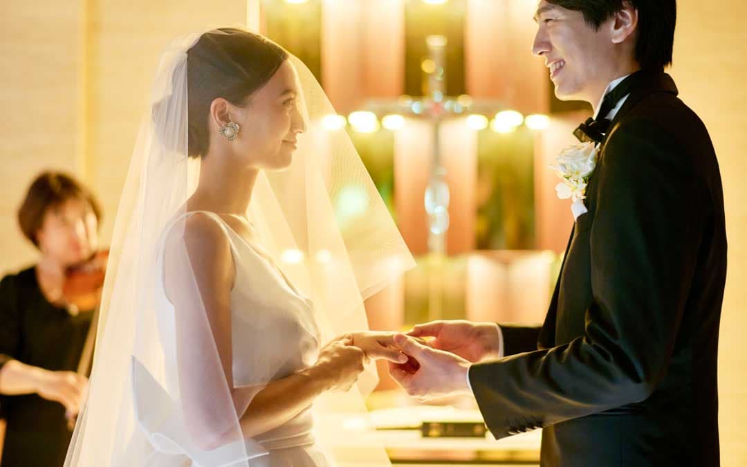 ウエディングチャペルで誓いの言葉を交わすカップル。新婦はベールをつけた白いドレスを着ており、新郎はタキシードを着ています。背景にはバイオリンを弾くセレモニーミュージシャンがいます。